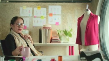 Asyalı orta yaşlı kadın moda tasarımcısı yaratıcı stüdyoda çalışıyor, çizim yapıyor, kahve içiyor, fikirler düşünüyor, elbise tasarımı koleksiyonunu görüntülüyor ve profesyonel butik terzisi KME.