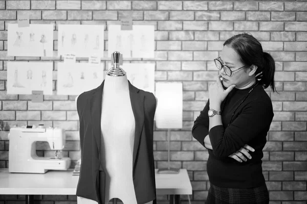 Black and White portrait, Asian mature female fashion designer stands near dressing puppet, thinking and contemplating for design ideas, clothing collection works, professional boutique tailor.