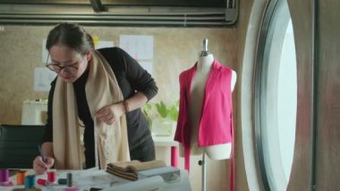 Asian middle-aged female fashion designer works in a studio by measuring puppet shapes and drawing sketches ideas, imagination dress design collection, professional boutique tailor SME entrepreneur.