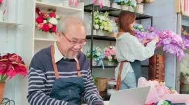 Asian elder male florist owner discussing with young beautiful female employee about floral bouquet arrangement and decoration for business, happy work in colorful flower shop store, e-commerce SME.