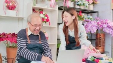 Asian elder male florist owner discussing with young beautiful female employee who shopkeeper about website arrangement for business service, happy work in colorful flower shop store, e-commerce SME. 