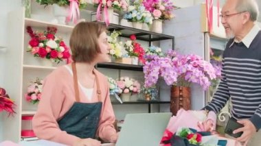 Cashless business entrepreneur. Asian elderly male customer shop and digital payments by scanning a mobile phone application to a White female florist owner. Beautiful floral shop, smart SME store.