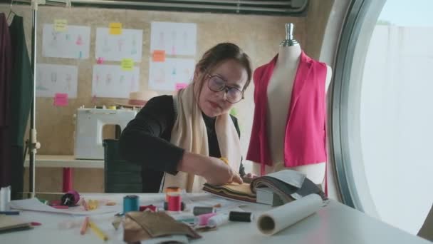 亚洲中年女性时装设计师在工作室工作 为服装设计系列裁剪和选择带有草图的面料和螺纹颜色 专业精品裁缝企业家 — 图库视频影像