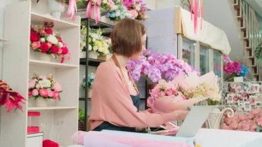One young Caucasian florist owner works with laptop, website arrangement for online business flora service in colorful flower shop store with beautiful bunch of blossom, e-commerce SME technology.