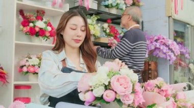 Young beautiful Asian female florist entrepreneur arranging a bunch of blossoms works with old man shopkeeper, happy work in colorful flower shop store with fresh blooms, small business, family SME. 