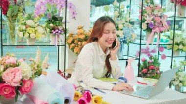 One young Asian female florist owner, working with laptop, selling floral arrange, talking on mobile phone in colorful flower shop store with a beautiful bunch of blossoms, and e-commerce business.