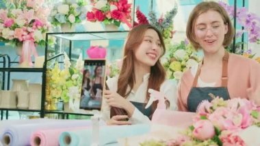 E-commerce business, two young female florist workers demonstrate and show floral arrangements via online live streaming with smartphone application in bright flower shop, a beautiful blossoms store.