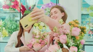 E-commerce business partner, two young female florist workers show floral arrangement, online live streaming, and selfies with smartphone application in bright flower shop, beautiful blossoms store. 