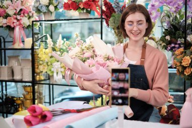 E-commerce business, one young White female florist demonstrates and shows floral arrangements via online live streaming with smartphone application in bright flower shop, beautiful blossoms store. 