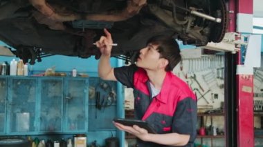 Professional young Asian male motor mechanic inspects undercarriage of electric car(EV) lifted by forklift jack for repair at garage, automotive maintenance service works industry occupation business.