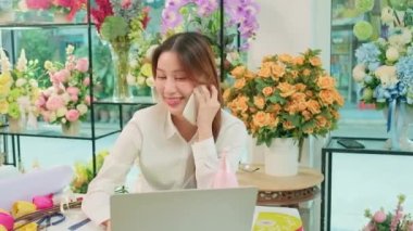 One young Asian female florist owner, working with laptop, selling floral arrange, talking on mobile phone in colorful flower shop store with a beautiful bunch of blossoms, and e-commerce business.
