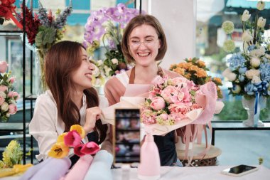 E-commerce business, two young female florist workers demonstrate and show floral arrangements via online live streaming with smartphone application in bright flower shop, a beautiful blossoms store.