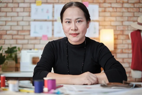 亚洲成熟的女性时装设计师正坐在桌旁 看着相机和工作室 与服装设计系列的创意和缝纫合作 专业精品裁缝店的中小企业创业者 图库图片