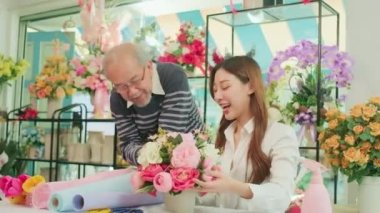 Asian young beautiful female florist worker and senior man shop owner arranging blossoms bouquet together in valentine, happy work with smile and cheerful in colorful flower store, SME entrepreneur. 