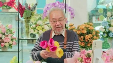 E-commerce business, one senior male florist workers demonstrate and show floral arrangements via online live streaming with smartphone application in a bright flower shop, a beautiful blossoms store. 