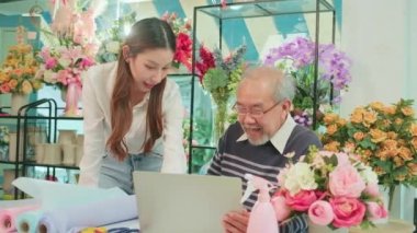 Asian elder male florist owner discussing with young beautiful female employee who shopkeeper about website arrangement for business service, happy work in colorful flower shop store, e-commerce SME. 