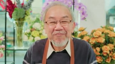 E-commerce business, one senior male florist worker shows floral arrangements and talks to camera via an online live-streaming webcam application in a bright flower shop, a beautiful blossoms store.