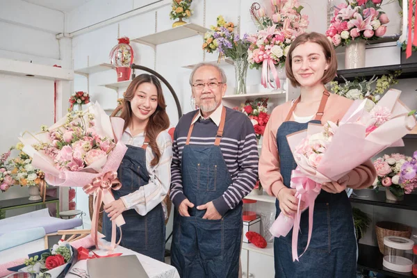 Portrait of flower shop workers team. Asian old male florist owner and young beautiful female employees in aprons with bunch of fresh floral bouquets arrangement for SME business, happy work in store.