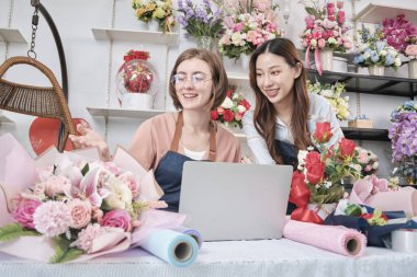Two young beautiful female florist partners discuss flora bunch decoration, online purchase orders and website arrangement for business, happy work in colorful flower shop store, and e-commerce SME.