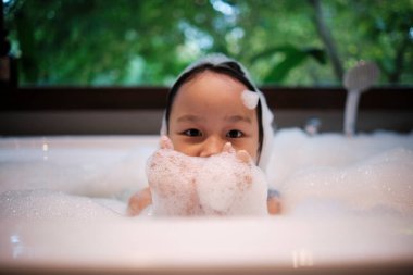 Asyalı şirin bir kızın portresi. Mutluluk ve eğlence dolu. Vücut sabunu köpüğü, şampuan sıçraması ve yeşil ağaç arka planlı bir otel banyosunda beyaz bir küvette su kabarcıkları..