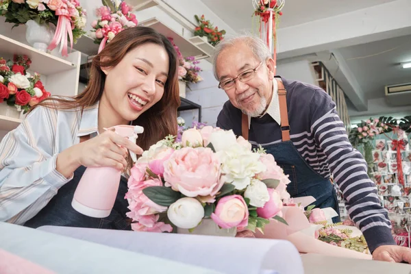 Old man and young beautiful Asian female florist workers water spraying a flora bunch together, happy work in a colorful flower shop store, fresh bloom bouquets decorating, SME business entrepreneur.