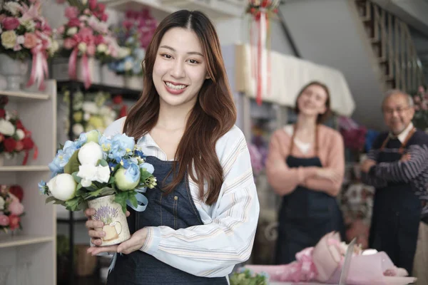 Beautiful young Asian female florist worker with vase of blossom smiles and looks at camera in front of colleagues team in colorful flower shop, small business occupation, happy SME entrepreneur.