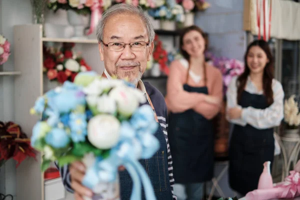 Senior Asian male florist owner in apron gives bunch of blossom smiles and looks at camera in front of her colleague\'s team in colorful flower shop, small business occupation, happy SME entrepreneur.