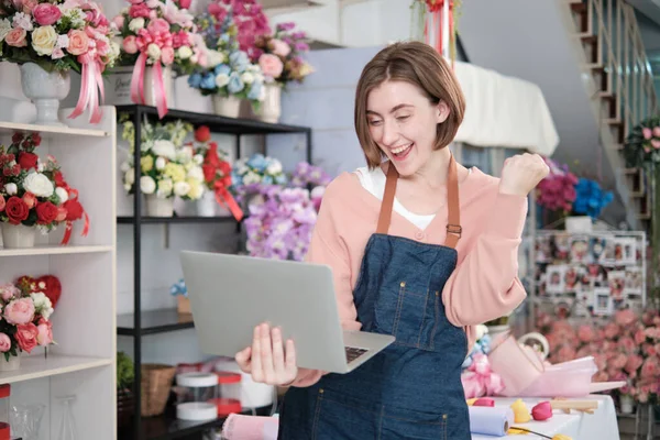 One young beautiful White female florist owner surprised and shocked purchase order profit, online income work in laptop at bright flower shop store, small e-commerce business, happy SME entrepreneur.