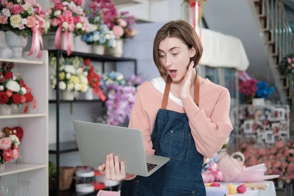 One young beautiful White female florist owner surprised and shocked purchase order profit, online income work in laptop at bright flower shop store, small e-commerce business, happy SME entrepreneur.