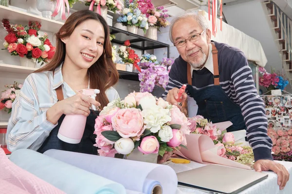 Old man and young beautiful Asian female florist workers water spraying a flora bunch together, happy work in a colorful flower shop store, fresh bloom bouquets decorating, SME business entrepreneur.