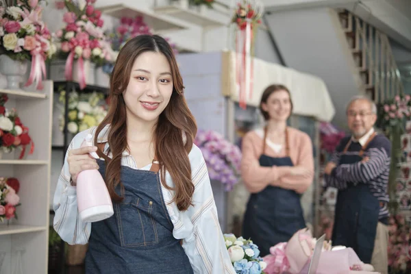 Beautiful young Asian female florist worker with water sprayer smiles and looks at camera in front of her colleague's team in colorful flower shop, small business occupation, happy SME entrepreneur.