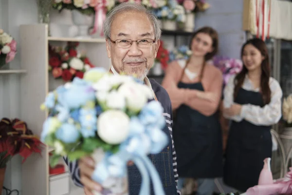 Senior Asian male florist owner in apron gives bunch of blossom smiles and looks at camera in front of her colleague\'s team in colorful flower shop, small business occupation, happy SME entrepreneur.