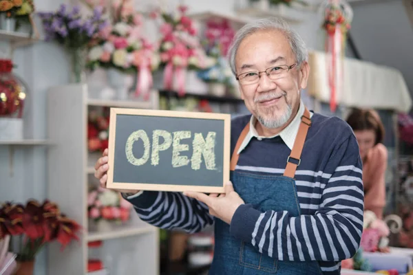 One senior male florist owner in apron shows open sign board in bright flower shop store with smile and looks at camera, small business opening, occupation retirement, happy elderly SME entrepreneur.