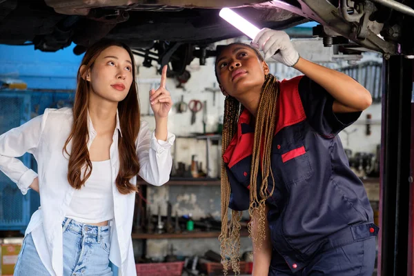 Young Asian female customer discusses with Black mechanical worker, check undercarriage of lifted electric car(EV) for repair at maintenance garage, automotive service center works industry business.