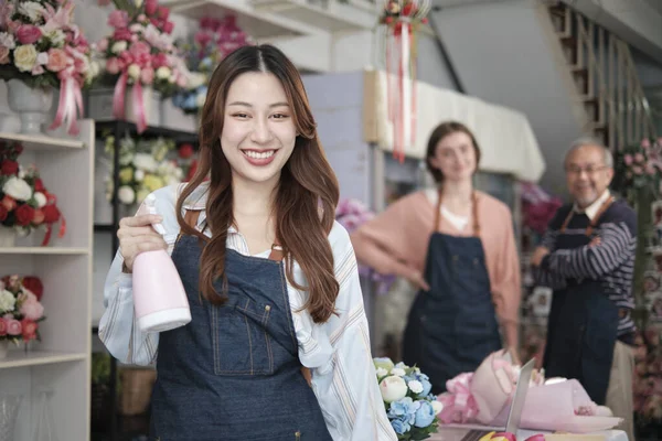 Beautiful young Asian female florist worker with water sprayer smiles and looks at camera in front of her colleague\'s team in colorful flower shop, small business occupation, happy SME entrepreneur.