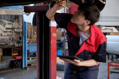 Profesyonel Asyalı genç erkek motor tamircisi, tamirat için forklift krikosuyla kaldırılan elektrikli arabanın (EV) bagajını teftiş ediyor..