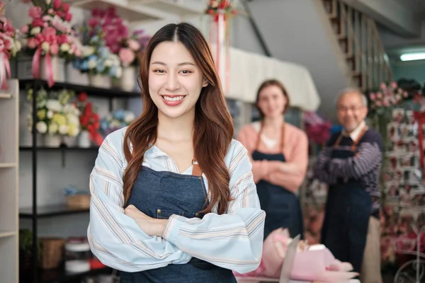 Beautiful young Asian female florist worker in apron, arms crossed, smiles and looks at camera in front of colleagues team in colorful flower shop, small business occupation, happy SME entrepreneur.