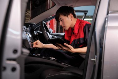 Genç bir Asyalı erkek otomotiv teknisyeni garajda içinde tablet bulunan bir bakım listesini kontrol ediyor. Araç servisi tamir ve onarım işleri, endüstriyel meslekler.
