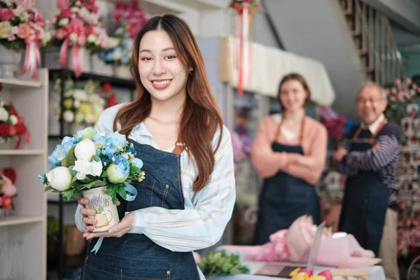 美丽的年轻亚洲女花匠 戴着花瓶 微笑着看着同事们面前的相机 在五颜六色的花店里 做小生意 快乐的中小企业创业者 图库图片