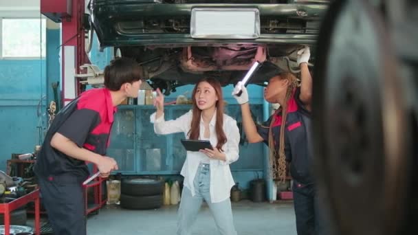 若いアジアの女性の自動車ビジネス起業家は 機械労働者チームと議論します メンテナンスガレージでの修理のためのエレベーター電気自動車のチェックリストの運送 自動車サービスの仕事業界 — ストック動画