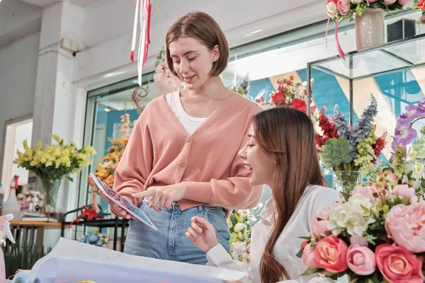 Two young beautiful female florist partners discuss bloom bouquet arrangement design in tablet, online purchase order work in colorful flower shop store with fresh flora, SME business entrepreneur.