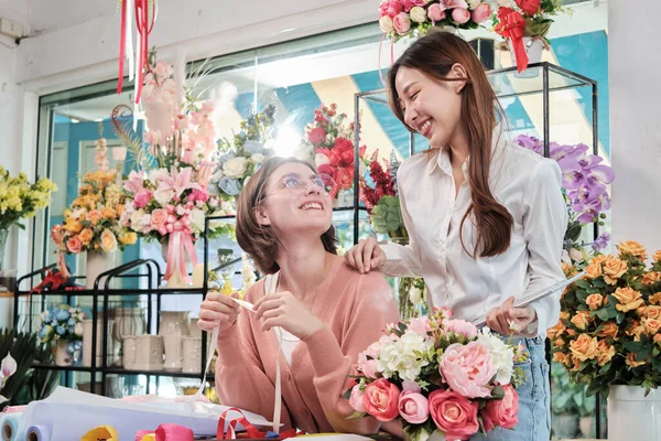 Portrait of two cheerful young beautiful female florist partners team smiling and looking together bloom bouquet happy work in colorful flower shop store with fresh flora, SME business entrepreneur.
