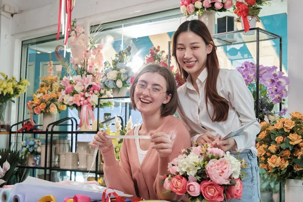 Portrait of two cheerful young beautiful female florist partners team smile and look at camera with bloom bouquet happy work in colorful flower shop store with fresh flora, SME business entrepreneur.