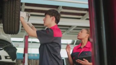 Genç uzman Asyalı erkek otomotiv tamircisi ingiliz anahtarıyla araba tekerleği taşaklarını beceriyor, tamirhanedeki kadın ortağın kontrol listesini kontrol ediyor. Araç bakımı sektör işlerinde çalışır.