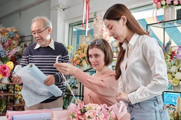 Flower shop team, Young beautiful female florist workers with old owner arranging and decorating bunch of fresh blossoms, smiling with happy work in colorful store, small business SME entrepreneur.