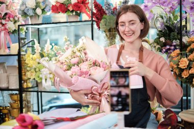 E-ticaret sektörü, genç beyaz kadın çiçekçi etkileyicisi çiçek aranjmanlarını gösteriyor ve gösteriyor, canlı yayında çiçekçide akıllı telefon uygulaması, güzel çiçek mağazası.