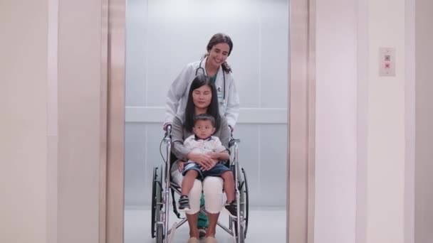 幸せな若い女性の小児科医は 外来診療所での診察後に車椅子で小さな男の子とお母さんを移動させるのに役立ちます 公共の健康診断 そして 予約訪問 — ストック動画