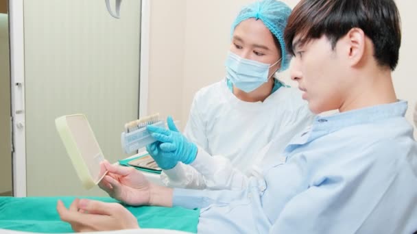 アジアの女性歯科医は 歯科医院 歯科衛生医師 病院の専門医療で歯の色合いを有する若い男性患者に矯正ケアの仕事とホワイトニングを説明します — ストック動画