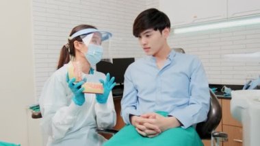 Asyalı kadın dişçi diş sağlığı kliniğinde diş modeli olan genç erkek hastaya diş bakımını ve beyazlatmayı, sağlık bakımını ve doktorluk hastanesinde profesyonel sağlık hizmetlerini anlatıyor..