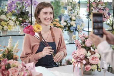 E-commerce business, one young White female florist demonstrates and shows floral arrangements via online live streaming with smartphone application in bright flower shop, beautiful blossoms store. 